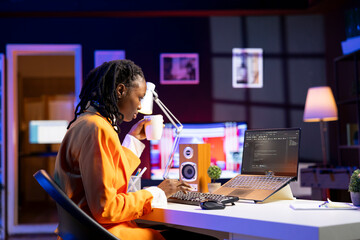Software developer enjoying cup of coffee and writing code on laptop in apartment office, using Java programming languages. Woman drinking beverage, remotely working on fixing database errors
