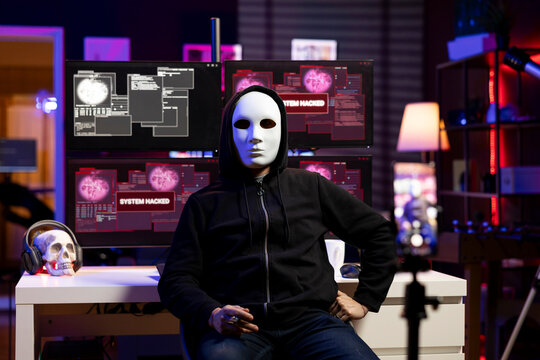 Evil programmer concealing identity using phone to film ransom video in secret base. Scammer with anonymous mask using smartphone to record blackmail footage, threatening victims
