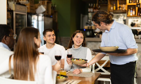 In restaurant, man waiter serves couple customer order, put plate with colorful salad of vegetables and cheese in front of guest. Cozy cafe establishment for family lunch, business meeting, coffee