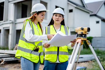 Two female engineers using theodolite for land survey at housing construction site, wearing reflective safety vests and hard hats while checking blueprints and project measurements