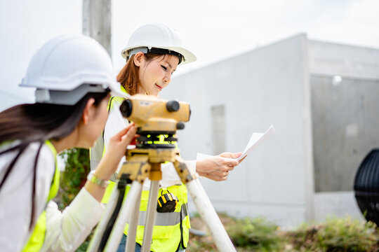 Two female engineers using theodolite for land survey at housing construction site, wearing reflective safety vests and hard hats while checking blueprints and project measurements