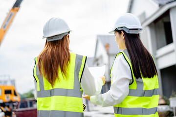 Back view of two female engineers wearing safety helmets and reflective vests reviewing construction blueprints and discussing project at building site, real estate developer, construction industry.