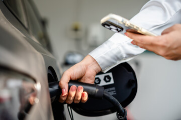 Close up of hand female holding smartphone while charging electric car at station. Concept of green energy, eco transport, smart technology and modern lifestyle with electric vehicles (EV).
