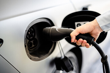 Close up of hand female holding smartphone while charging electric car at station. Concept of green energy, eco transport, smart technology and modern lifestyle with electric vehicles (EV).