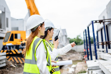 Two female engineers wearing safety vests and helmets discussing construction blueprints at residential building site with crane in background, teamwork and project planning concept
