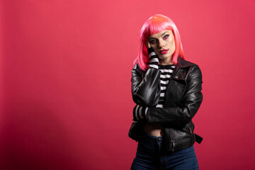 Beautiful pink haired woman poses with hand on cheek, wearing leather jacket and striped shirt, exuding bold elegance in studio. Female model with vibrant hair and calm expression poses gracefully.