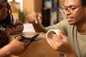 Black woman with scissors assisting boyfriend with cutting clear tape, sealing cardboard box of belongings during moving day. African american couple packing together, getting ready to move out.