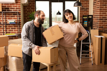 Portrait of caucasian couple carrying belongings in bright living room. Husband and wife embracing new beginnings and joyful relocation, woman holding box with hand on waist while man looks on.