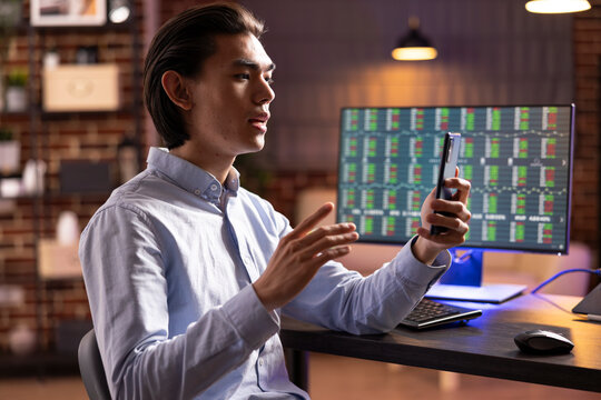 Serious business consultant communicates with clients on smartphone, giving trading updates from computer. Asian man manages freelance business remotely, discussing ideas and plans with global