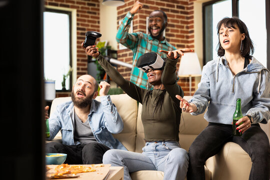 Multiethnic group of friends enjoy augmented reality and online video games in cozy apartment. African American woman with VR glasses reacts to gameplay as diverse peers hold drinks and socialize. - Powered by Adobe