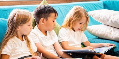 Three children reading on a couch. Two girls and one boy, all in school uniforms, focused on a...