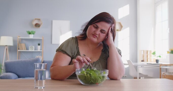 Depressed fat plus-size woman reluctantly eating a vegetable salad at home. Struggling with obesity and embracing healthy food choices, her expression reflects feelings of inner conflict.