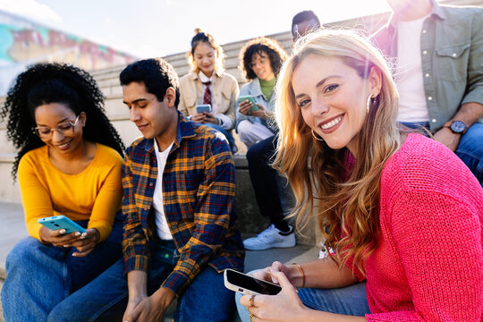 Group of multiracial students gathered outdoors sharing a smartphone, while one young woman looking at the camera. Youth community and friendship concept. - Powered by Adobe