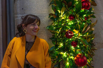 Profile portrait of a woman in yellow coat and gold earrings, looking at an elegant Christmas decoration of bright green leaves, lights and an orange sphere. Festive atmosphere at night.