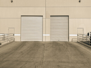 Two large metal roll up doors on an industrial warehouse building with a concrete loading dock and railings under bright daylight in a quiet urban business area