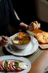 A person enjoys a bowl of flavorful Georgian soup filled with fresh vegetables and herbs, while a platter of vibrant tomato and basil salad sits nearby