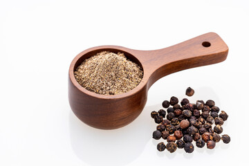 Ground black pepper in a spoon surrounded by whole peppercorns on a white background