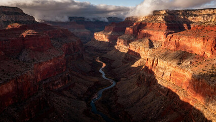 Obraz premium Aerial view of a deep canyon with layered rock formations and a winding river below, illuminated by sunlight under a partly cloudy sky.