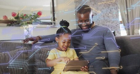 Guiding father leaning beside girl in yellow dress at home, using tablet showing holograms