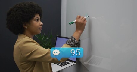 Writing woman in tan blazer holding laptop at office whiteboard, using green marker and 95 chat