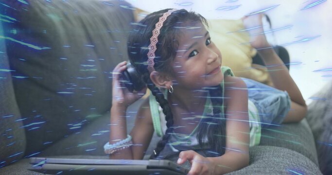 Lying girl in tank top and denim shorts on grey couch, holding tablet, pressing over-ear headphone