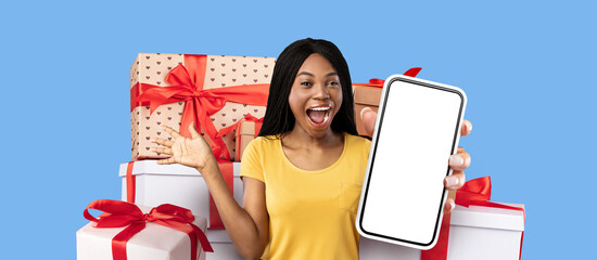 A joyful woman holds a smartphone while standing in front of a collection of beautifully wrapped gifts. She expresses excitement and happiness, celebrating a special moment.