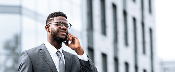 A well-dressed man stands outside a contemporary office building, engaged in a phone conversation while smiling. The scene captures a professional moment in a bustling city.