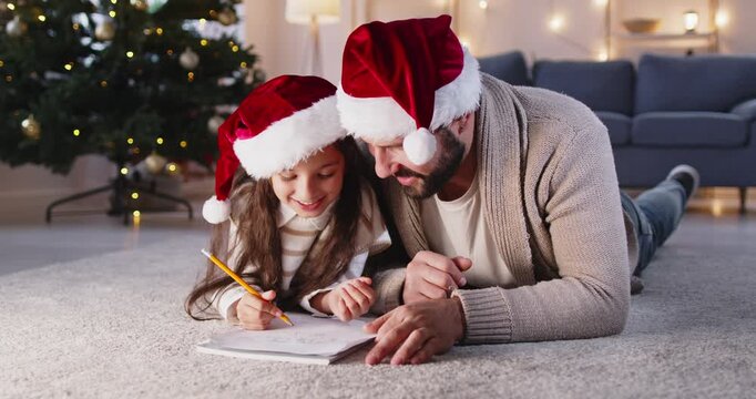 Happy father and daughter write Christmas letter or draw in notebook together at home. Child in Santa hat thinking and writing wish list with dads help, people lying by Xmas tree under holiday lights