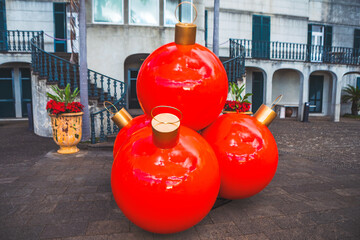 Oversized red Christmas ornaments with gold caps sit clustered on paved plaza, reflecting light. Vibrant red holiday decorations contrasting with muted tones of exterior building facade in background
