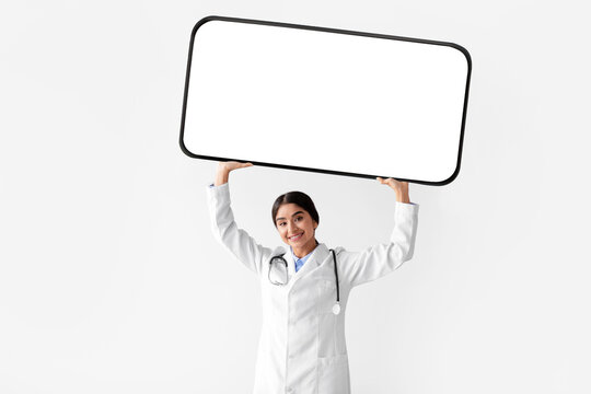 A happy young Hindu female in a white coat raises a large smartphone with a blank screen above her head. She is promoting remote doctor consultations and new medical offers in a bright, clean space.