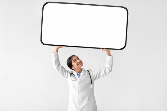 A happy young Hindu woman doctor in a white coat holds a big phone with a blank screen above her head. She stands against a light background, representing modern healthcare and digital solutions.