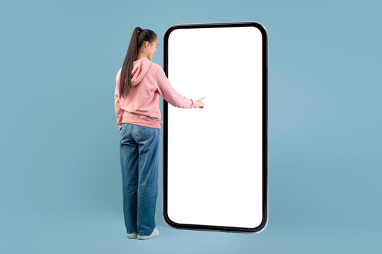 A young woman with long hair in a casual outfit smiles while touching the empty display of a huge smartphone. She is engaged in exploring a new app against a blue background.