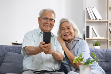 Asian Senior couple selfie happy grandparents smiling elders smartphone living room casual clothing silver hair glasses cozy home togetherness