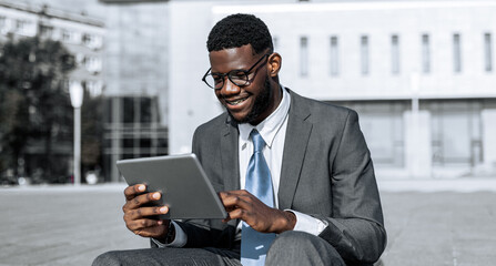 A young man in a suit and tie sits on the ground outside a contemporary building, focused on his tablet. He looks pleased and engaged, enjoying his time in a vibrant urban space.