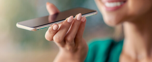 A woman holds her smartphone to her ear while smiling. She appears to be making a voice call outdoors in a pleasant and sunny environment.
