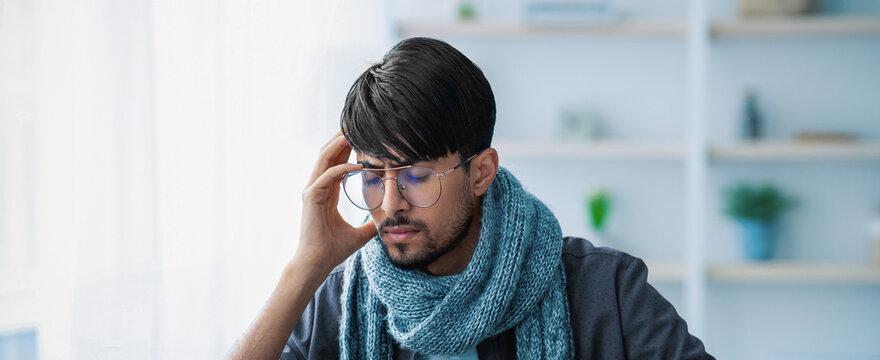 A young man with stylish glasses and a blue scarf looks pensive while seated at a table in a bright, modern indoor environment. He appears to be deep in thought, possibly contemplating ideas or tasks.