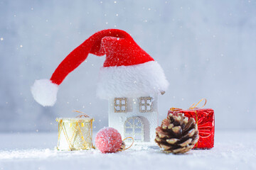 Small decorative Christmas house wearing a Santa hat surrounded by a gift box, pine cone and ornaments in snow. Festive winter holiday concept for Christmas greeting cards and New Year marketing.
