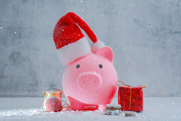 Pink piggy bank with Santa hat stands in a snowy holiday setting with Christmas decorations and gift boxes. Festive winter composition for savings, financial planning and holiday spending.
