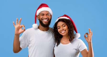 Xmas time. Excited african american spouses showing OK gestures, wearing Santa hats, posing and...