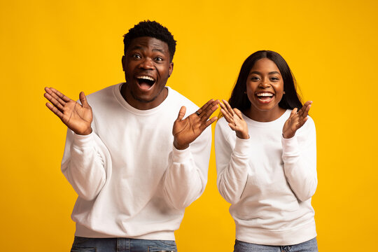 Two friends express happiness and excitement with wide smiles and playful gestures. They are wearing matching casual sweaters and denim jeans, set against a bright yellow backdrop.