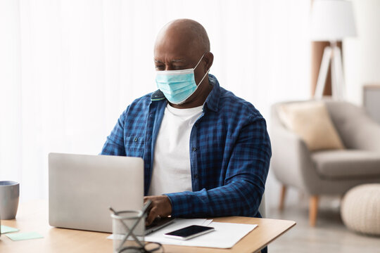 Senior African American businessman focuses on his laptop in a bright office space while wearing a face mask. This scene highlights remote work and employee safety during the pandemic.
