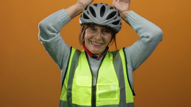 Senior hispanic woman wearing a helmet and safety vest smiling against an orange background, making playful ear gestures above her head, conveying a sense of humor and safety.