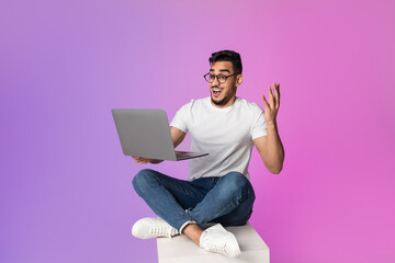 Excited young Arab man sits cross-legged with a laptop, feeling overjoyed by a significant online sale. He expresses surprise and happiness in bright neon lighting.