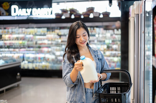 Fototapeta Asian woman holding shopping basket buying or choosing fresh milk gallon from fridge in supermarket.