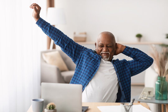Senior African American man sits at his desk in a modern office, stretching his arms after a successful day. He enjoys a moment of relaxation in a peaceful work environment. - Powered by Adobe
