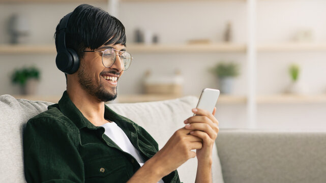 A young man wears headphones and smiles as he checks his smartphone in a comfortable living room. The space is bright and decorated with plants, creating a relaxed atmosphere.