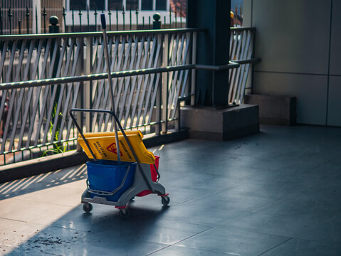 Cleaning trolley with mop bucket and caution sign placed on a tiled floor near railing, symbolizing hygiene, maintenance, and cleanliness in public or commercial areas.