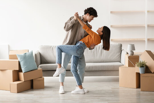 A joyful young Asian couple dances closely in their living room among unpacked cardboard boxes on moving day. They share a moment of happiness as they embrace their new beginning.