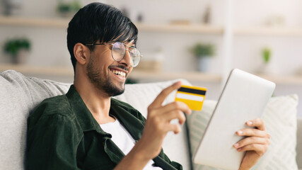 A young man smiles as he holds a credit card in one hand and uses a tablet in the other while relaxing on a couch. He appears engaged in online shopping in a cozy home environment.