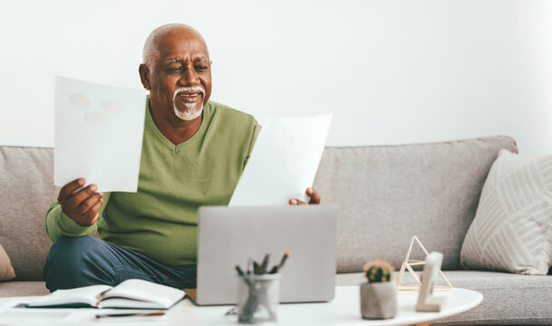 A senior man sits comfortably on his sofa, holding papers while looking at his laptop. He appears engaged and happy in a bright, modern living room with minimal decor. - Powered by Adobe